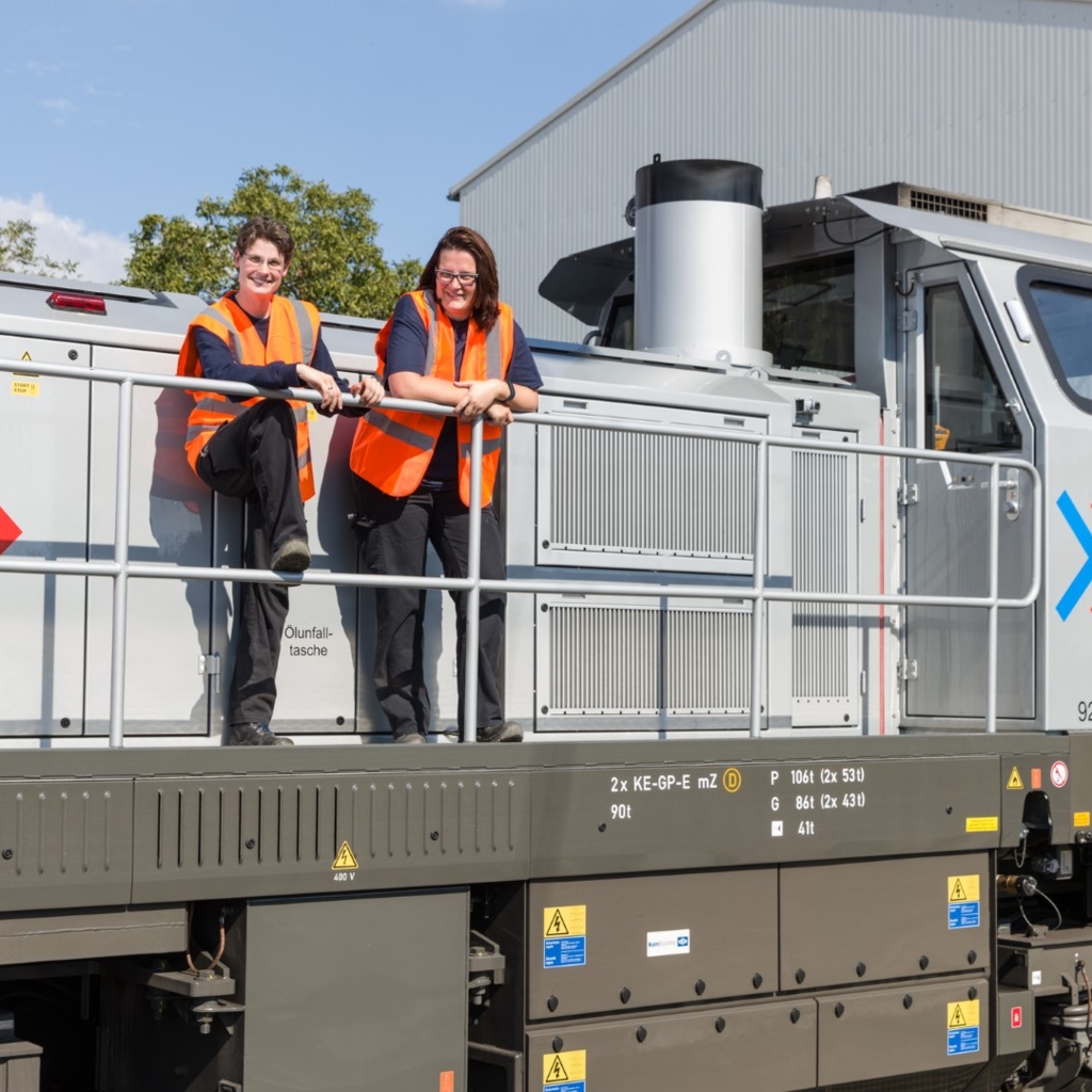 Zwei RheinCargo-Mitarbeitende tragen orangefarbene Warnwesten und stehen auf der Plattform einer silbergrauen Lokomotive mit der Aufschrift „DE 502 RheinCargo“. Sie lächeln in die Kamera, im Hintergrund sind ein Industriegebäude, Bäume und blauer Himmel zu sehen.