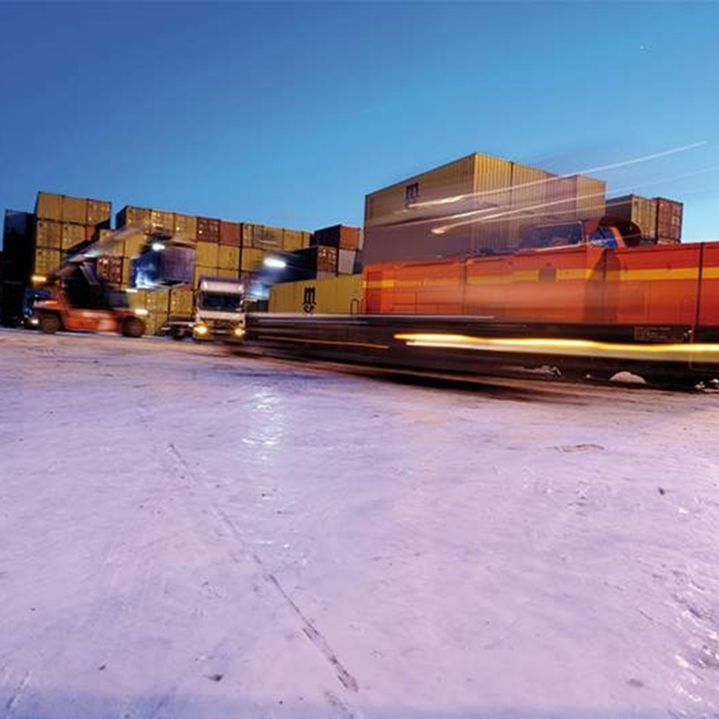 Eine rote Diesellok bewegt sich mit hoher Geschwindigkeit über das verschneite Gelände eines Containerterminals. Im Hintergrund sind gestapelte Container, Kräne und Lkw im Einsatz. Die Szene ist in der Dämmerung aufgenommen, das Licht der Scheinwerfer und der hell erleuchtete Himmel erzeugen eine dynamische, industrielle Atmosphäre.