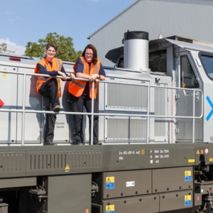 Zwei RheinCargo-Mitarbeitende tragen orangefarbene Warnwesten und stehen auf der Plattform einer silbergrauen Lokomotive mit der Aufschrift „DE 502 RheinCargo“. Sie lächeln in die Kamera, im Hintergrund sind ein Industriegebäude, Bäume und blauer Himmel zu sehen.